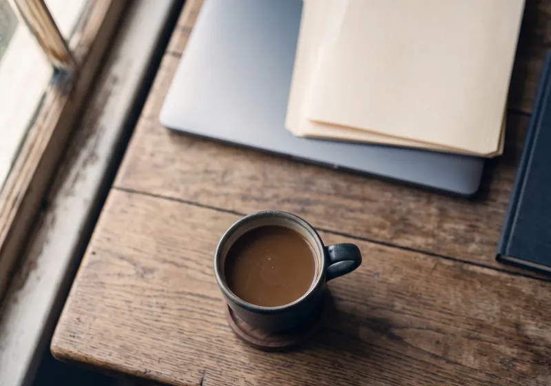Taza de café junto a un portátil cerrado y una carpeta de documentos sobre una mesa de madera.